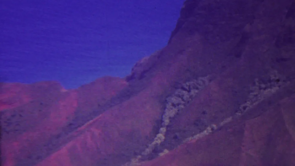 1962 Hawaii 8mm frame showing a dark cliff face dropping toward reddish-brown terrain, pale vegetation or exposed stone, and blue coastal water in the background.
