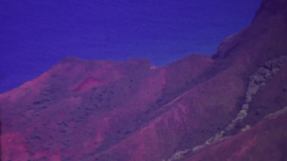 1962 Hawaii 8mm frame looking across reddish rugged ground toward a steep cliff edge and blue ocean or distant water, with purple-blue aged-film color throughout.