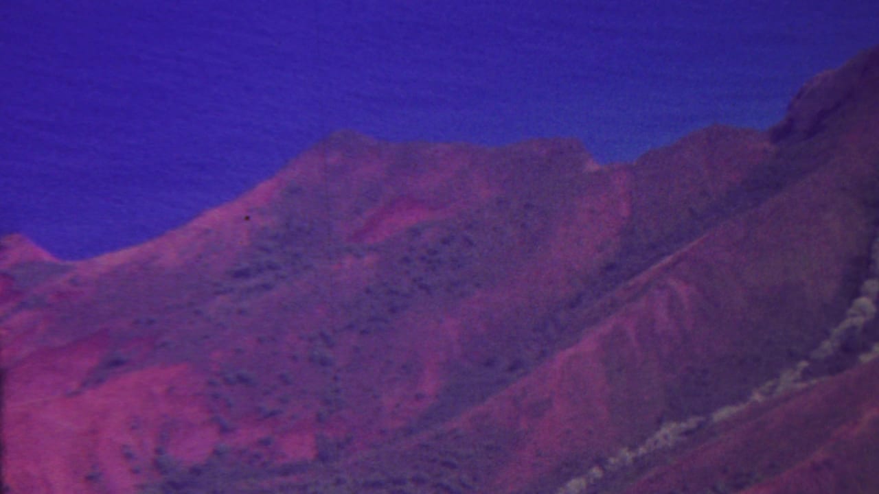 1962 8mm Hawaii footage showing a reddish-purple mountain ridge under a saturated blue sky, with green shrubs and trees along the lower slope.