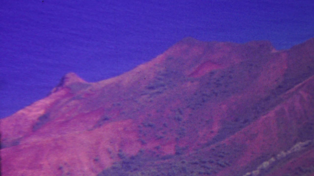 1962 8mm Hawaii footage with a rugged red mountain peak, darker ridge shadows, and a clear saturated blue sky.