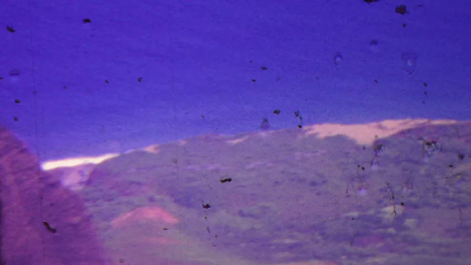1962 8mm Hawaii footage showing a distant green-and-red mountain slope beneath blue sky, with visible dust specks and exposure variation from the film scan.