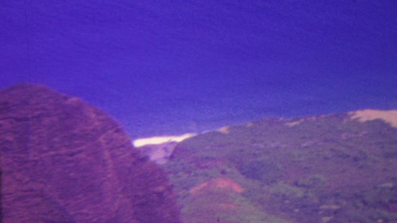 1962 Hawaii 8mm frame showing a hazy mountain ridge with green slopes under a strong purple-blue sky and a bright strip of distant ocean or surf.
