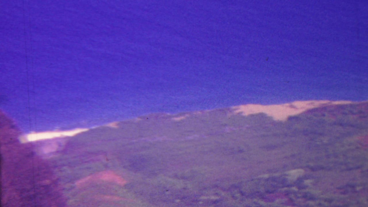 1962 Hawaii home-movie frame with a soft green mountain ridge, purple-shifted sky, and a pale horizontal line of distant water or surf behind the landform.