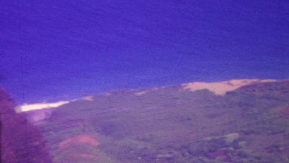 1962 Hawaii home-movie frame with a soft green mountain ridge, purple-shifted sky, and a pale horizontal line of distant water or surf behind the landform.