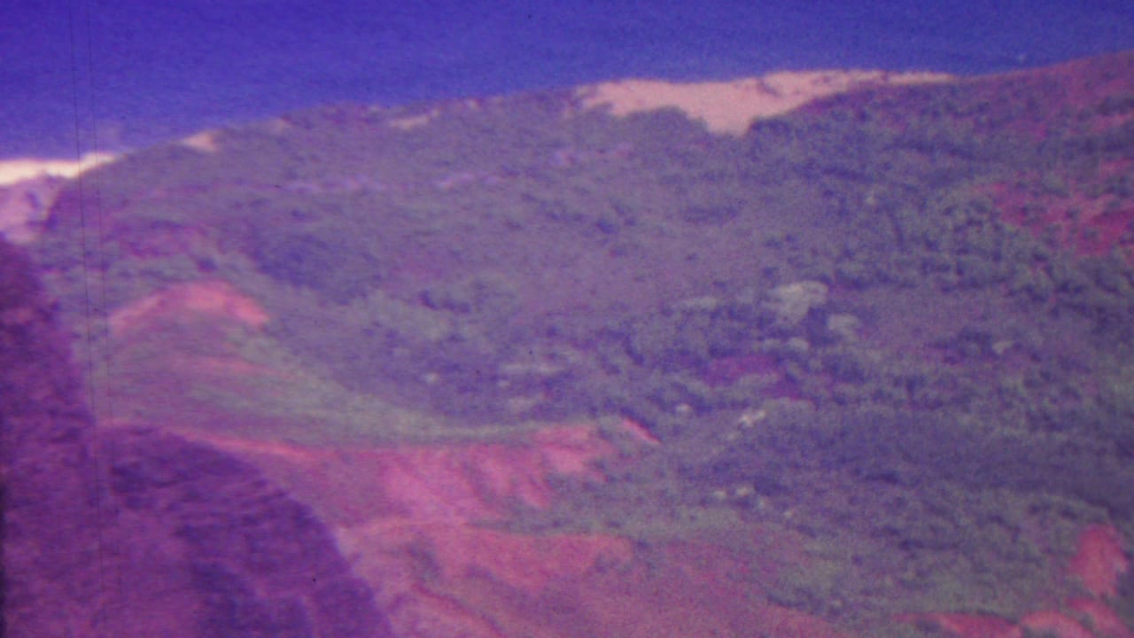 1962 Hawaii 8mm aerial frame showing rolling green terrain, red soil patches, distant ridges, and aged magenta color shift.