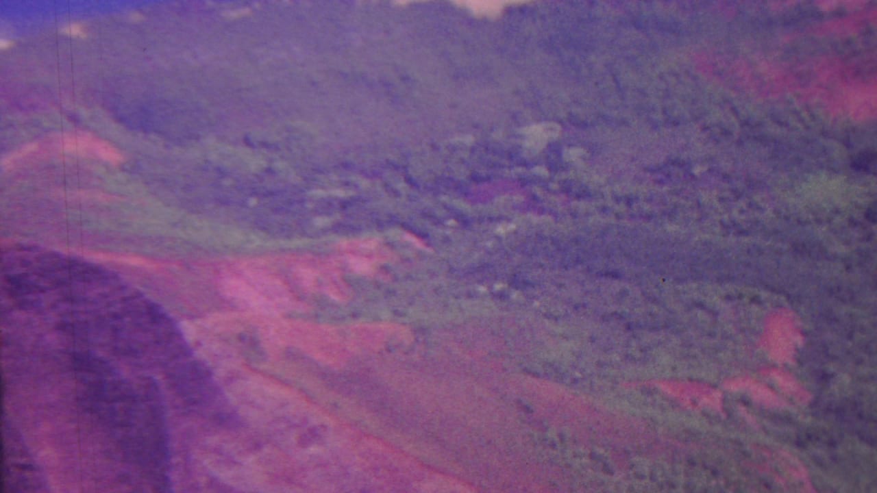 1962 Hawaii aerial home-movie frame looking across rugged vegetation, valley folds, red soil, and ridgelines with pronounced color fading.