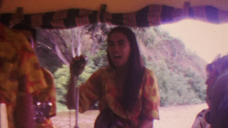 1962 Hawaii 8mm frame showing a female performer singing near a microphone under a striped boat canopy with water and foliage behind her.
