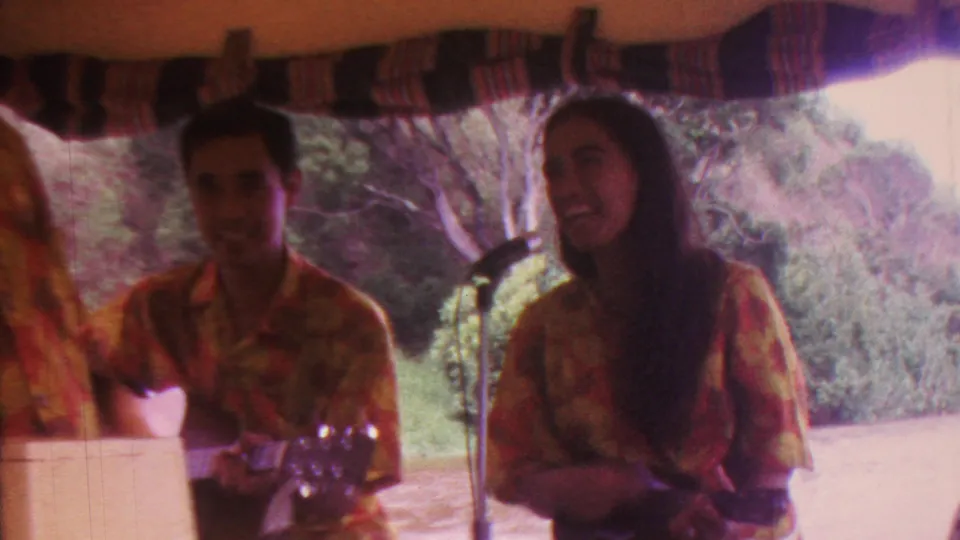 1962 Hawaii boat performance frame with a male musician and female ukulele player smiling beneath a canopy, with tropical foliage in the background.