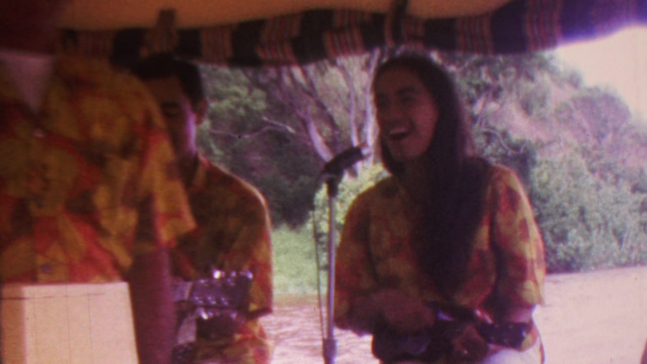 In metadata-supported 1962 Hawaii footage, a woman in a yellow and red floral shirt sings into a microphone while holding a ukulele or small stringed instrument beside another performer.