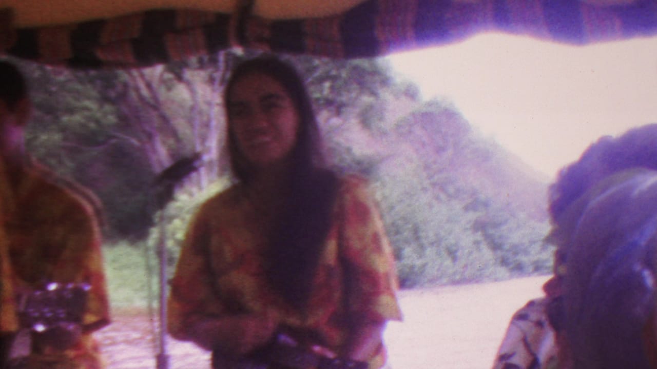 Woman performer faces forward near a microphone with trees and hillside behind her.