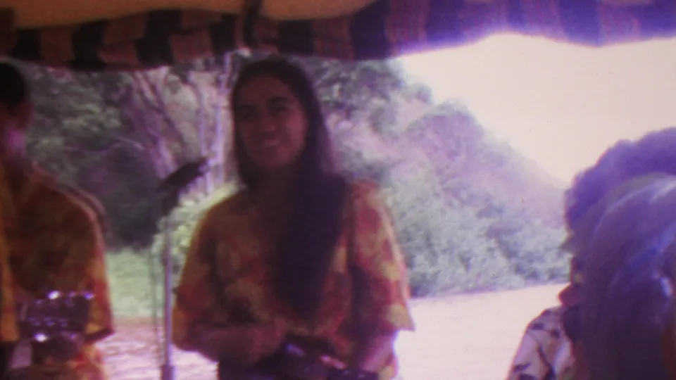 A woman performer in a floral shirt stands near a microphone under a canopy in the 1962 Hawaii home-movie clip, with a bright outdoor landscape behind her.