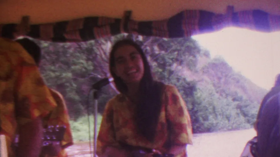 The smiling woman singer holds a ukulele or similar small stringed instrument near the microphone, with the canopy edge and green hillside visible in the 1962 Hawaii setting.