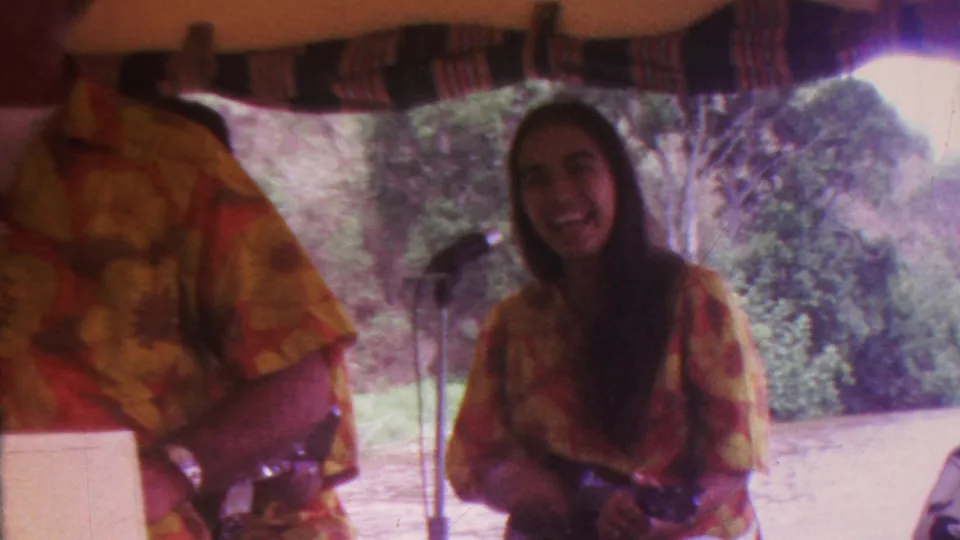 A smiling woman performer stands beside the microphone while another musician in a matching floral shirt partially blocks the foreground, with trees and hillside behind the outdoor stage area.