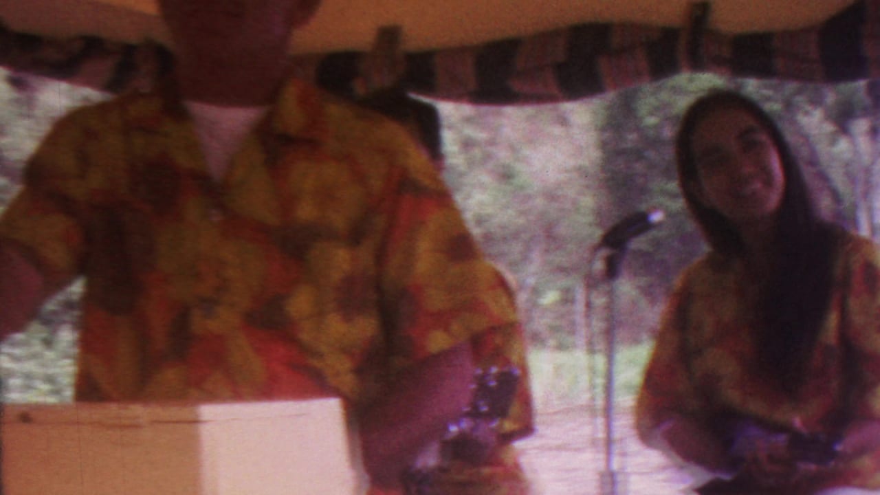 In Hawaii home-movie footage identified as 1962, adults in yellow-red patterned shirts stand under a covered pavilion near a lectern and microphone, with trees visible outside.