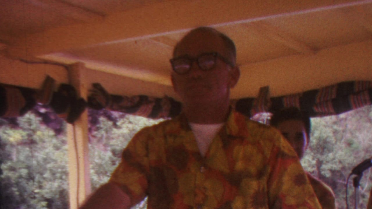 A man wearing glasses and a yellow-red patterned shirt appears inside a covered pavilion in the Hawaii footage, with another person partly visible and leafy trees beyond the windows.