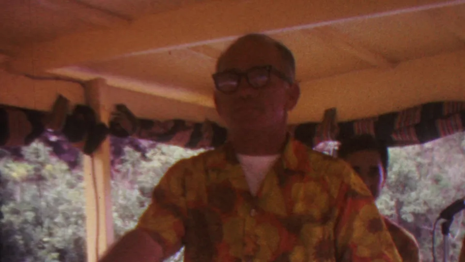 A man wearing glasses and a yellow-red patterned shirt appears inside a covered pavilion in the Hawaii footage, with another person partly visible and leafy trees beyond the windows.