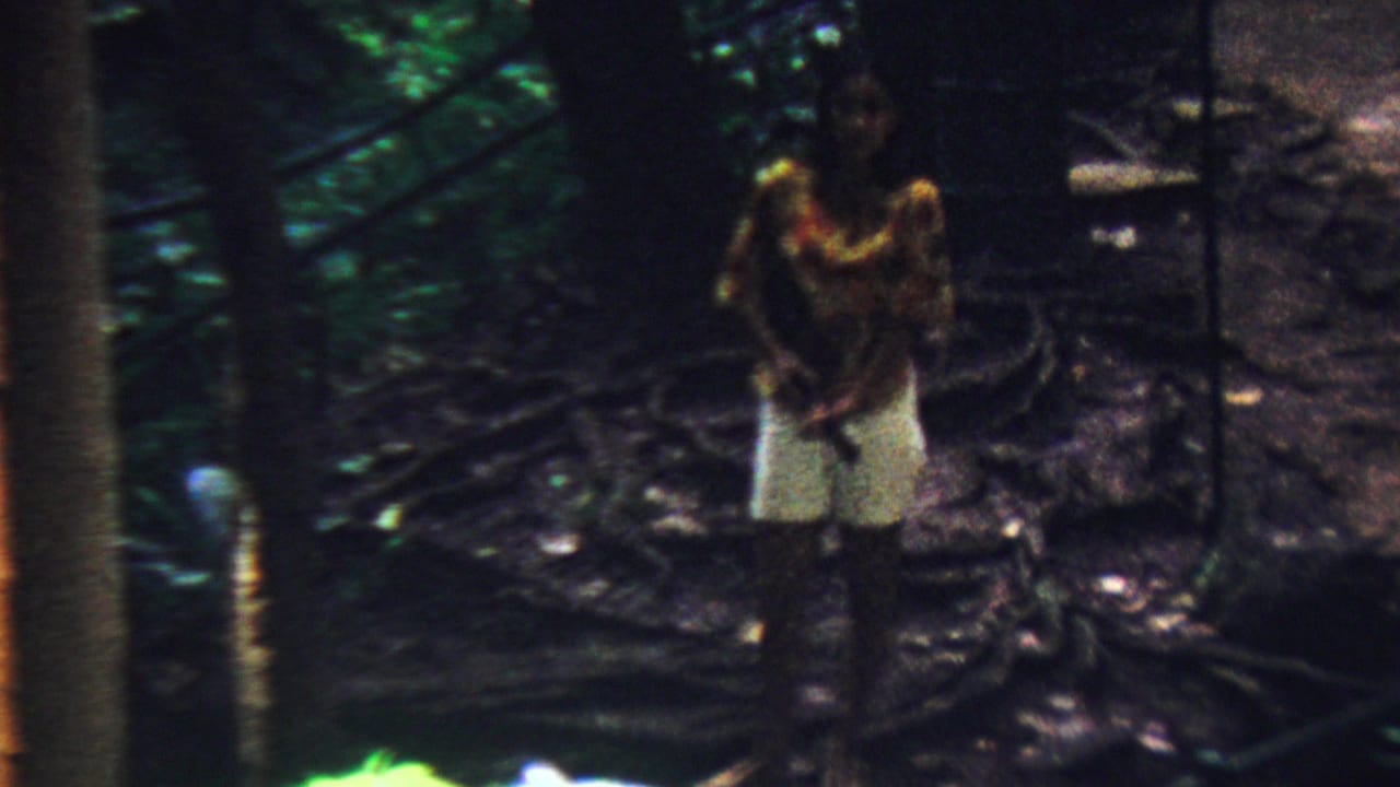 The woman remains near tree trunks and dark tropical foliage, with her bright shirt and white shorts visible in the soft 8mm frame.