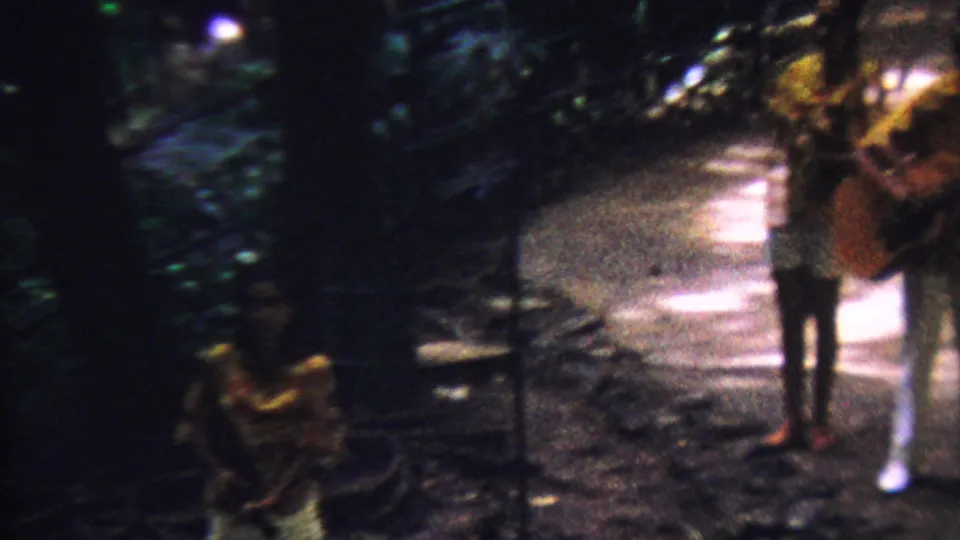 Two adult musicians, one partly obscured, stand with acoustic guitars on a dappled tropical path in the 1962 Hawaii home-movie scene.