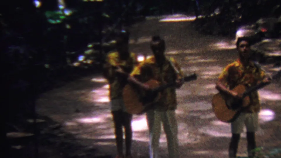 Three adult musicians, including the woman in a patterned shirt, hold acoustic guitars together on a shaded tropical path in Hawaii footage dated 1962.