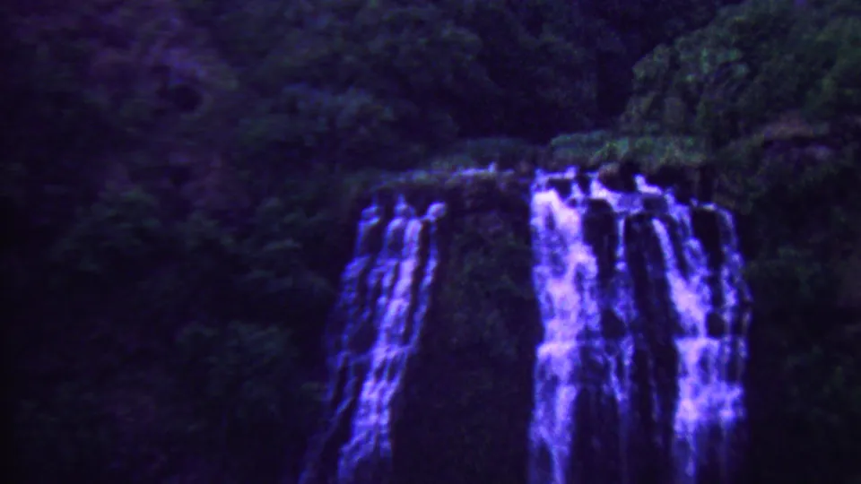 A closer 8mm view shows multiple streams of waterfall dropping over dark rock through tropical greenery in Hawaii.
