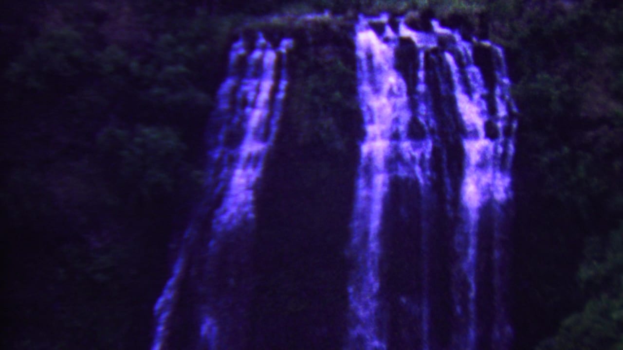 1962 Hawaii 8mm frame showing several blue-purple waterfall streams dropping over dark rock with dense green forest around them.