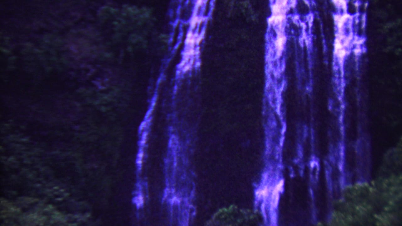 1962 Hawaii 8mm frame with tall blue-purple waterfall curtains beside shadowed tropical vegetation and a dark cliff face.