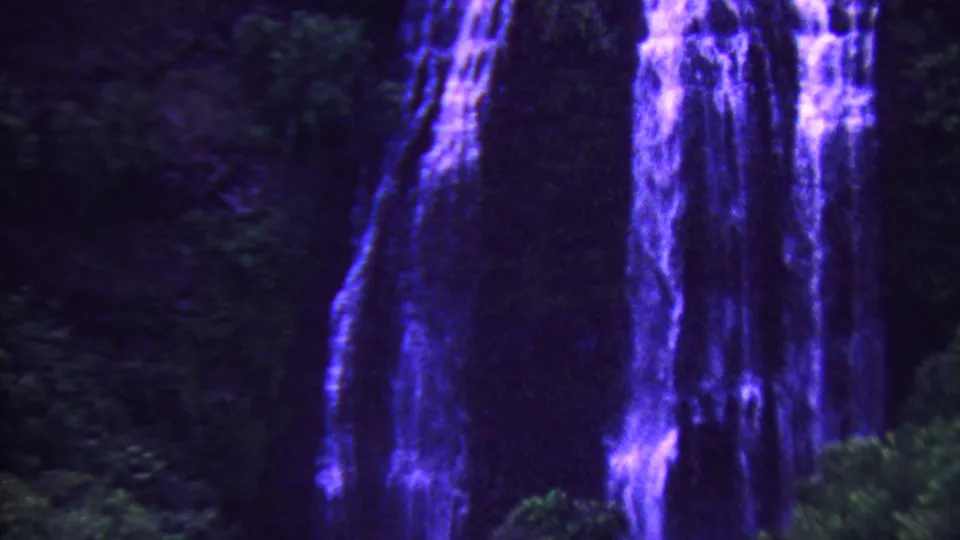 1962 Hawaii 8mm frame with tall blue-purple waterfall curtains beside shadowed tropical vegetation and a dark cliff face.