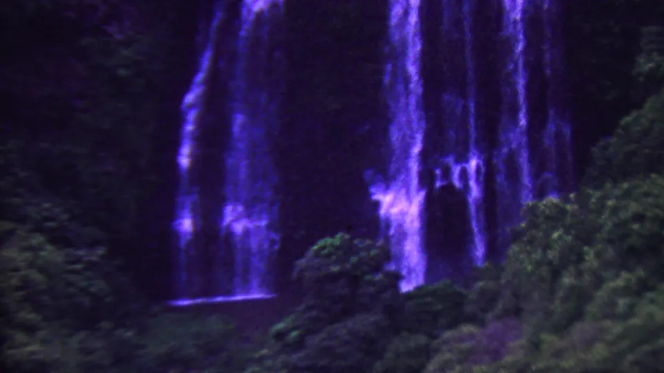 1962 Hawaii 8mm frame showing a wider view of cascading blue-purple water, rock ledges, and dense tropical forest near the base of the falls.