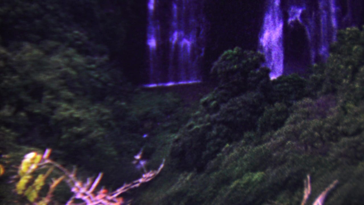 1962 8mm Hawaii home-movie frame showing purple-tinted waterfall streams partly obscured by dense tropical greenery.