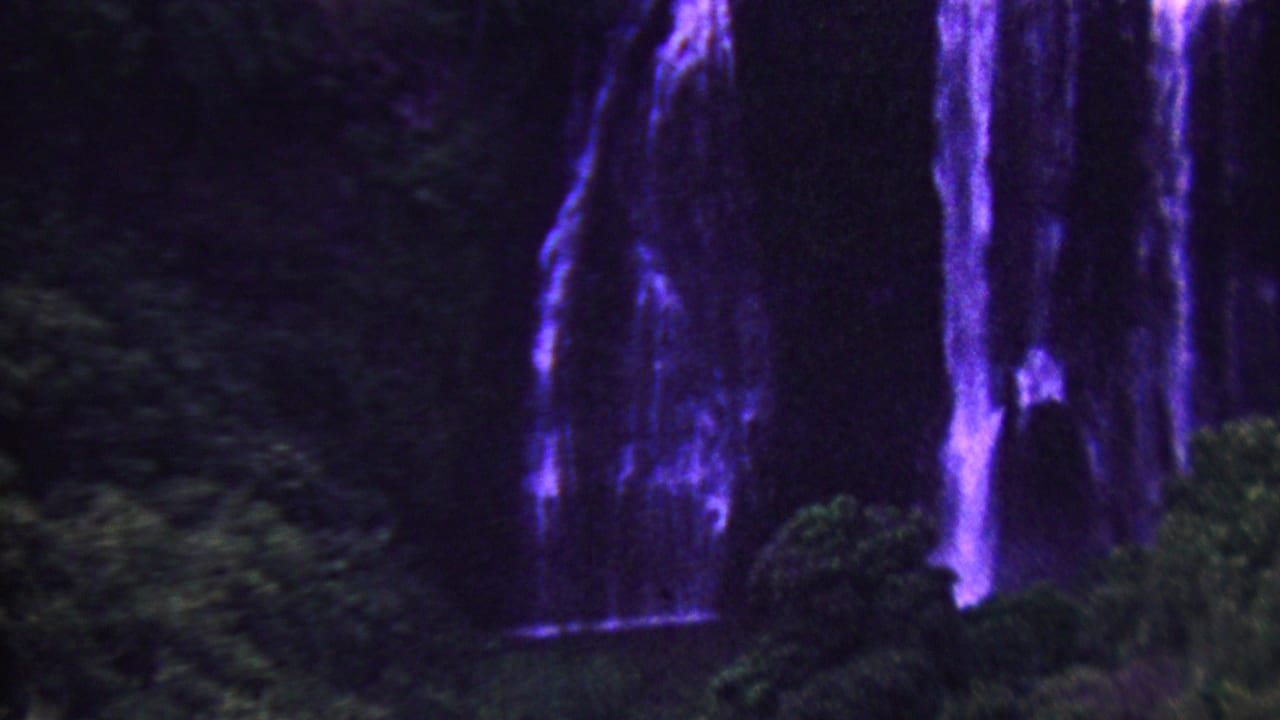 1962 Hawaii travel footage frame with a tall purple-blue waterfall descending over a dark cliff amid tropical vegetation.