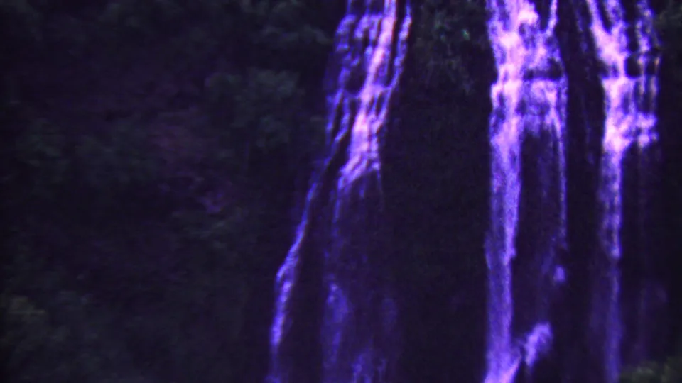 Close 8mm frame of a purple-tinted Hawaiian waterfall falling down a dark, heavily shadowed rock wall with foliage at the edges.
