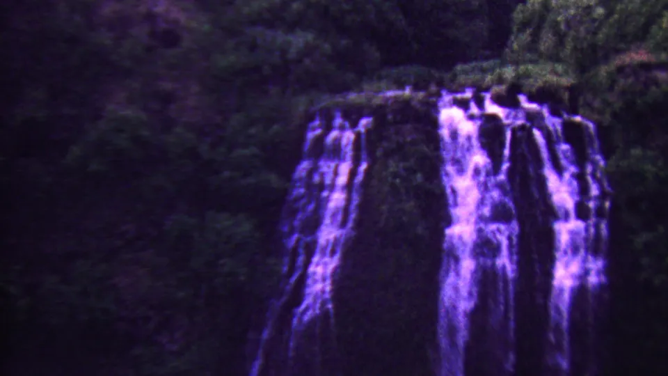 1962 8mm Hawaii frame showing multiple purple-tinted cascades beside leafy vegetation on a shadowed forest slope.