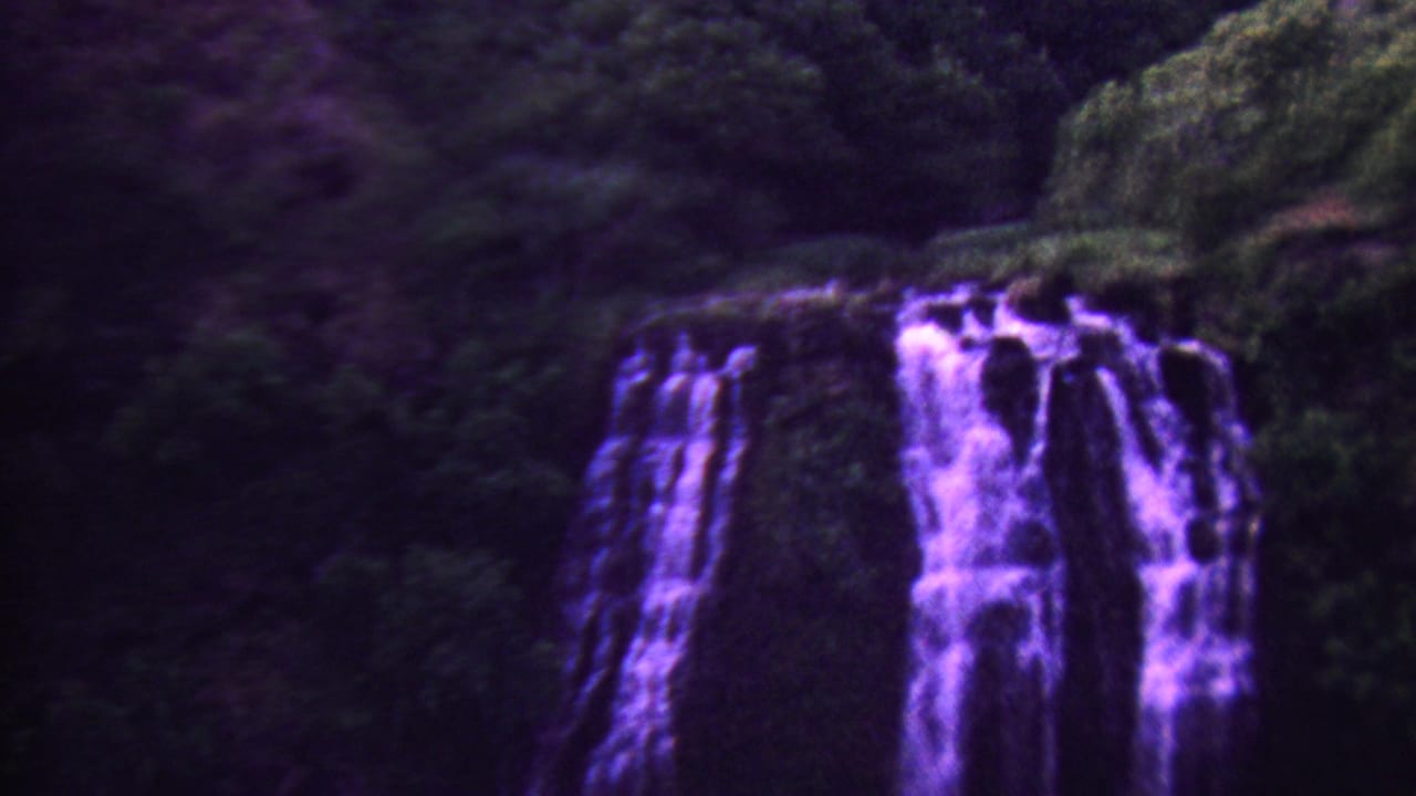 1962 Hawaii 8mm frame showing a shaded waterfall surrounded by dense green vegetation, rendered with pronounced magenta-purple film saturation.