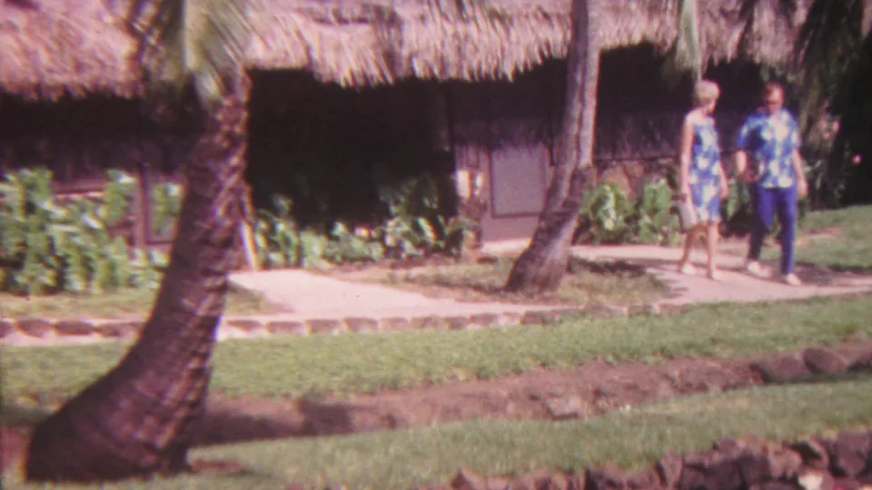 1962 Hawaii home-movie frame showing two adult visitors walking along a paved garden path near a thatched-roof building, palms, lawn, and tropical plantings.