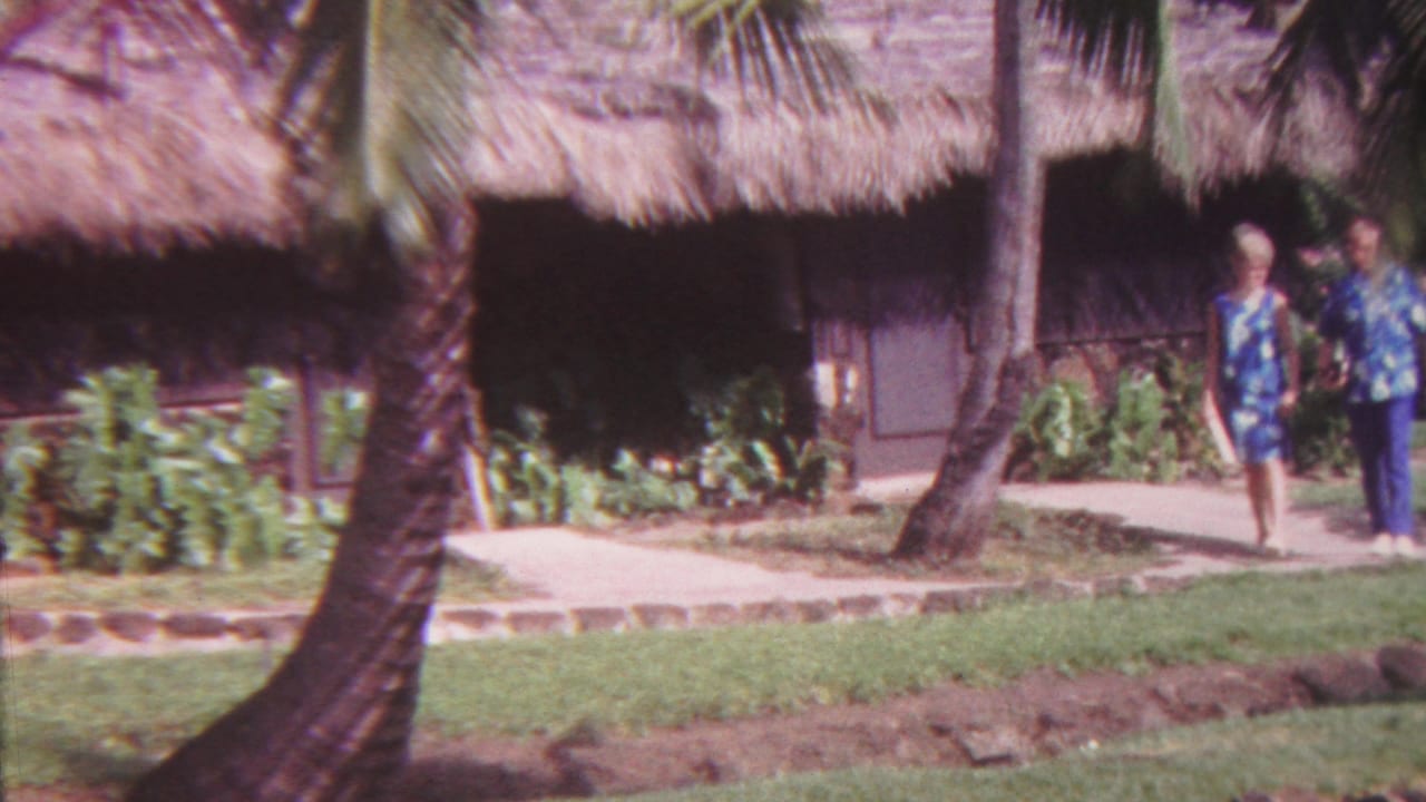 In 1962 Hawaii home-movie footage, an adult couple in Hawaiian-print clothing walks along a paved tropical path beside palms and a thatched-roof structure.