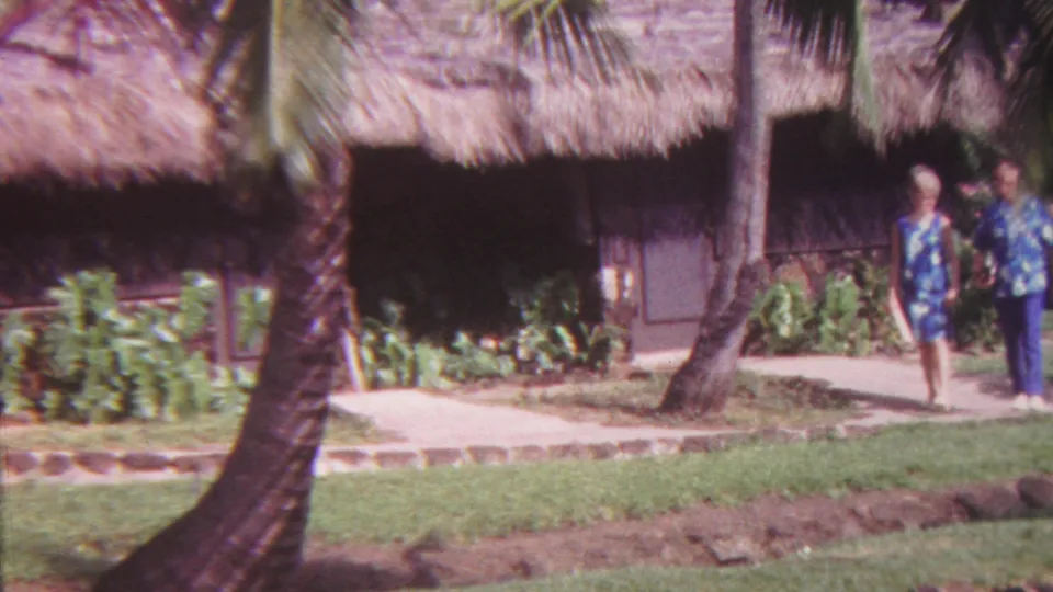In 1962 Hawaii home-movie footage, an adult couple in Hawaiian-print clothing walks along a paved tropical path beside palms and a thatched-roof structure.