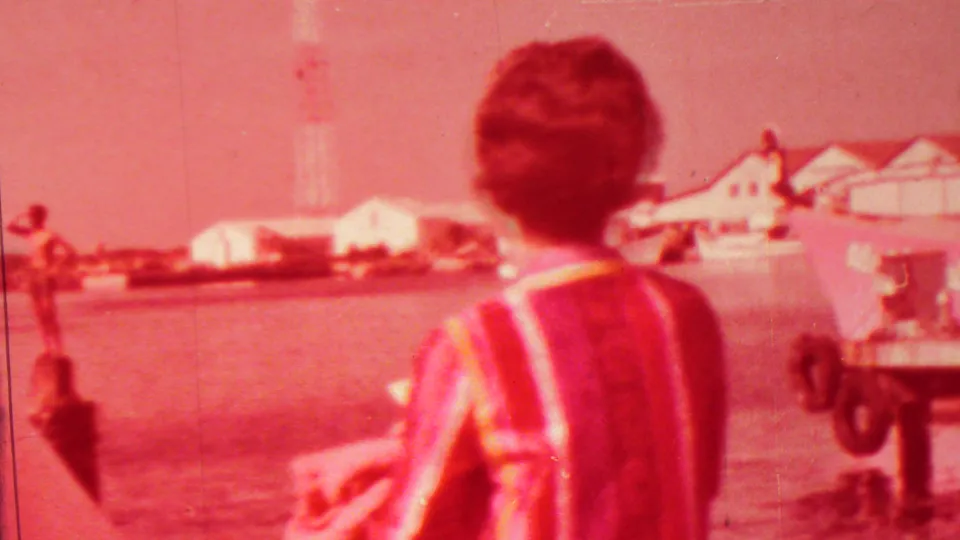 Man in a striped shirt viewed from behind as he looks across Pearl Harbor water toward boats, low buildings, and a tall structure in 1962 Hawaii footage.