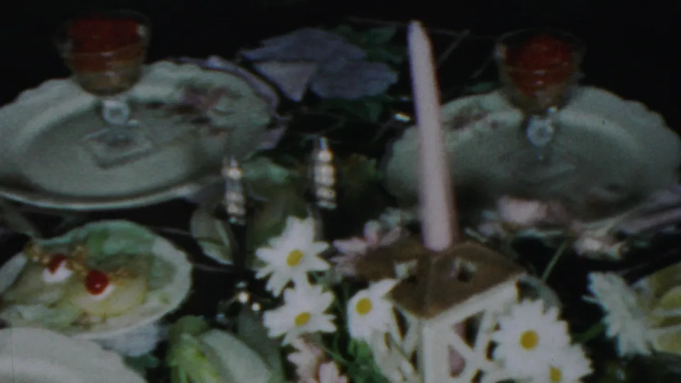 1954 Columbus, Ohio home-movie frame showing a formal tea table with china plates, crystal-style glasses, white daisies, molded desserts, and a candle lantern centerpiece.