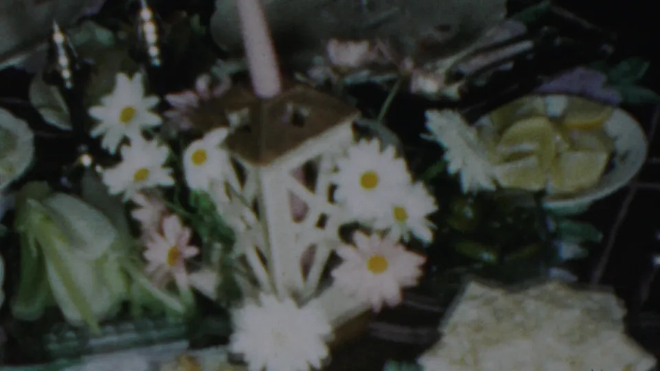 1954 Columbus, Ohio home-movie frame with white daisies around a candle lantern centerpiece, china plates, glassware, and arranged desserts on a formal table.
