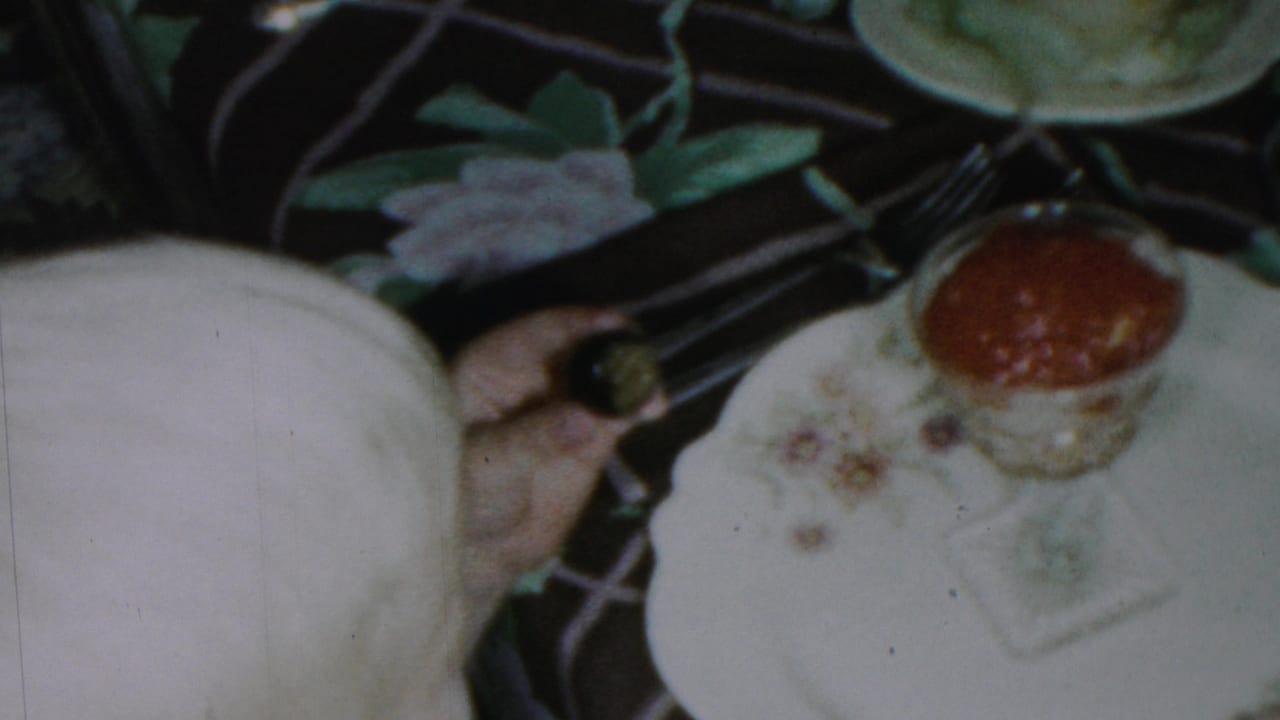1954 Columbus, Ohio home-movie frame showing a formal table setting with floral plates, a glass of red sauce, and serving dishes on a dark patterned tablecloth.