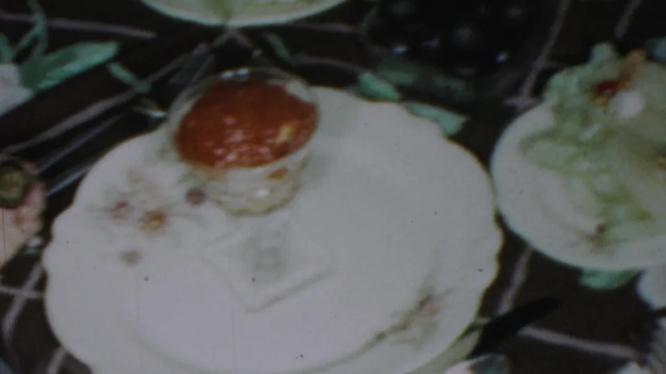 1954 Columbus, Ohio home-movie frame with a floral-patterned plate, glass of red sauce, and surrounding dishes in a formal dining arrangement.