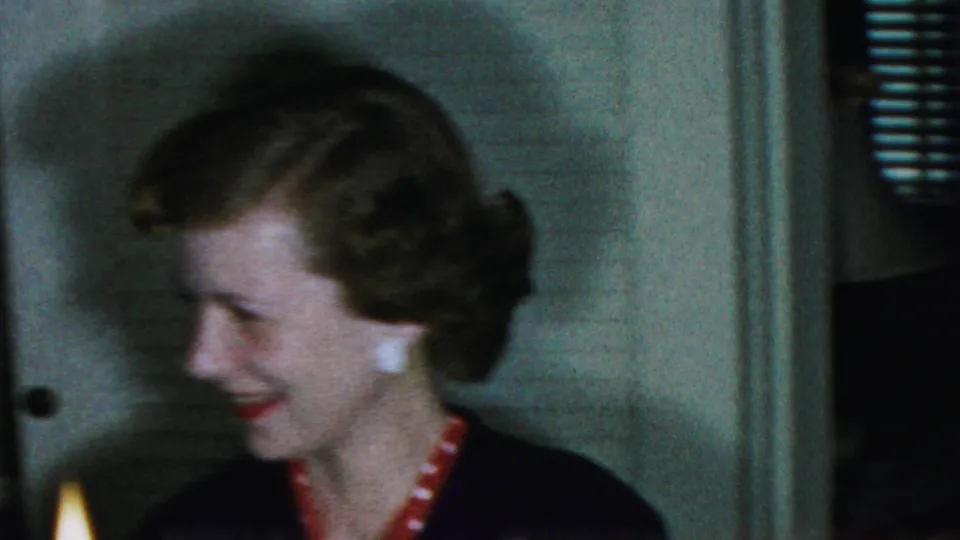 In the 1954 Columbus, Ohio home-movie scene, the woman smiles toward someone off to the side while candlelight appears at the lower edge of the indoor frame.