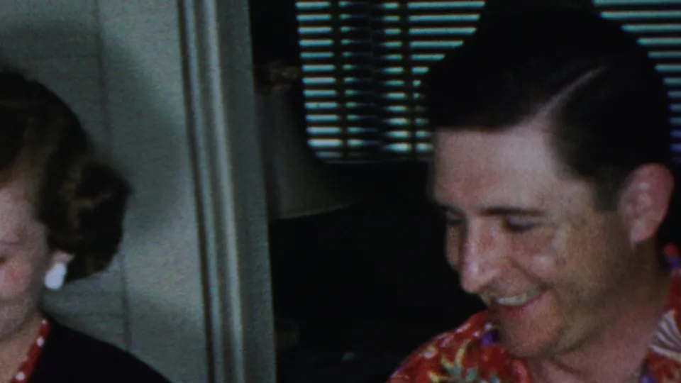A man in a colorful tropical-print shirt laughs inside the same private home setting, with part of the woman visible at the left and venetian blinds behind him.