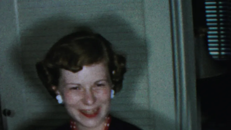 The woman smiles directly in the indoor 1954 Columbus, Ohio home-movie frame, with soft focus, a wall panel, and venetian blinds visible behind her.