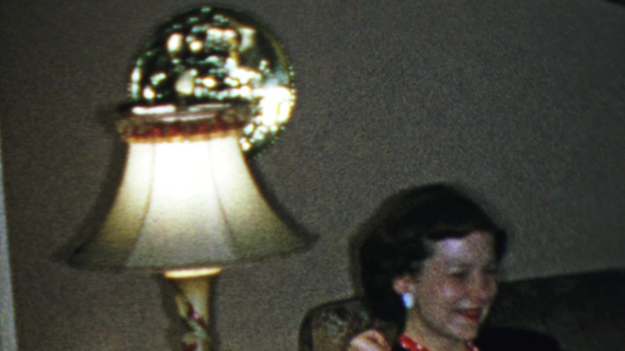 In a 1954 Columbus, Ohio home-movie frame, a well-dressed woman sits near an upholstered chair or couch beside a bright table lamp in a dim living room.