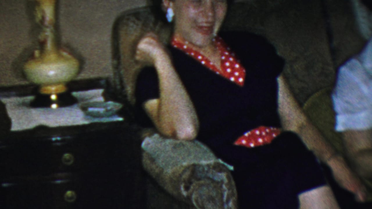 In supplied 1954 Columbus home-movie footage, a young woman in a dark dress with red polka-dot trim laughs on a patterned living-room sofa near a decorative lamp.