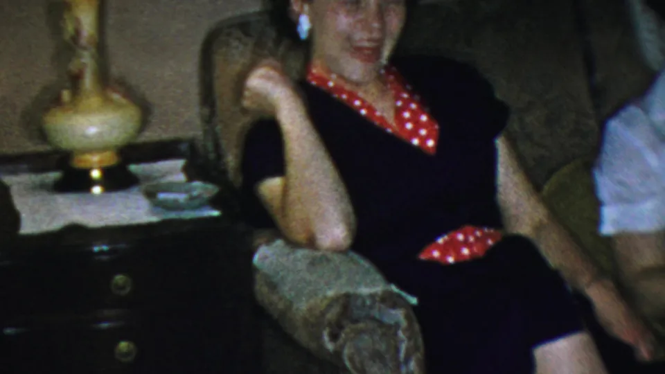 In supplied 1954 Columbus home-movie footage, a young woman in a dark dress with red polka-dot trim laughs on a patterned living-room sofa near a decorative lamp.