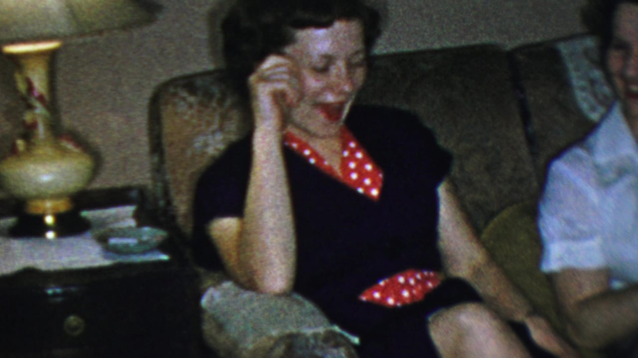 Woman in a dark dress laughs with one hand raised near her face while seated on a sofa.