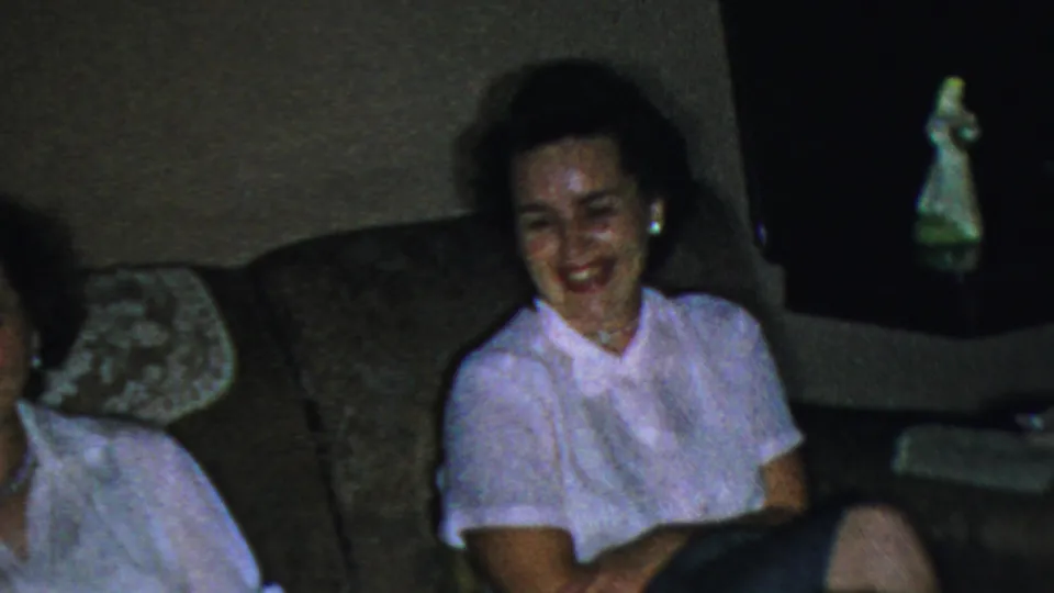 In Columbus, Ohio home-movie footage from 1954, an adult woman in a light pink short-sleeved collared shirt laughs while seated on a dark couch in a dim living room.