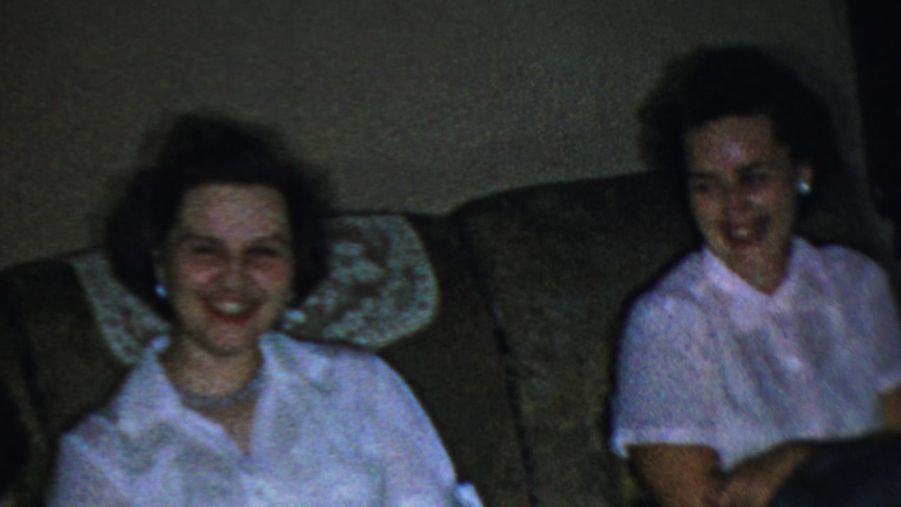 Woman in a white blouse smiles from a couch beside another partially visible seated woman.